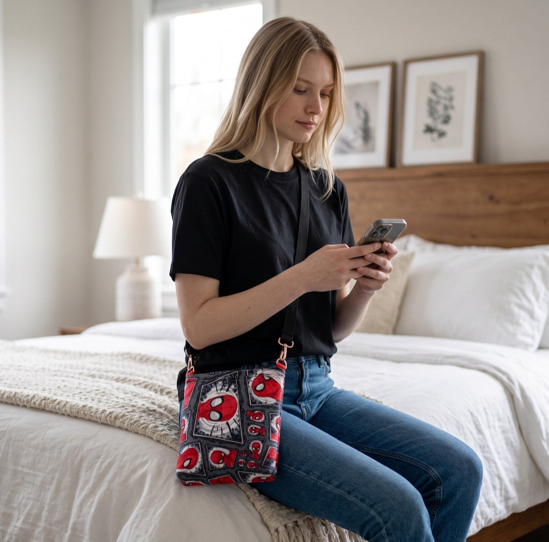 A woman wearing a JUST-JEN BOUTIQUE Spider-Man graphic print crossbody bag while sitting on a bed and using her phone.